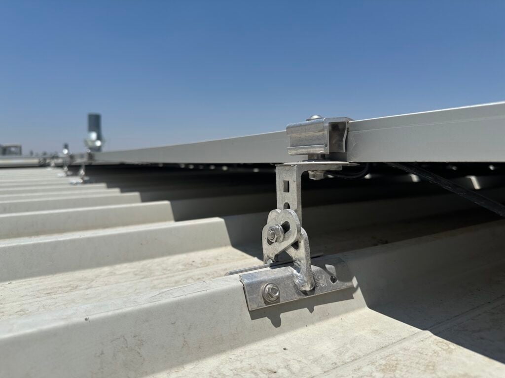 Vista lateral de un sistema de montaje de panel solar sobre un techo de lámina trapezoidal. La estructura metálica está fijada al techo con pernos, sosteniendo un riel de aluminio en el que va instalado el panel. El cielo está despejado y azul.
