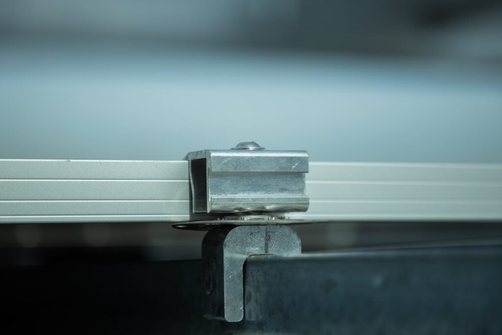 Vista lateral en primer plano de una abrazadera metálica que sujeta un riel de aluminio, como parte de un sistema de montaje de paneles solares. La abrazadera está atornillada a un techo de metal corrugado y se distingue ligeramente la marca S-5!.